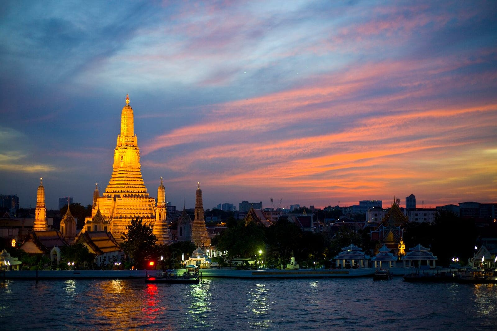 Wat Arun temple in Bangkok lit up at sunset, with the Chao Phraya River in the foreground and a pink and orange sky