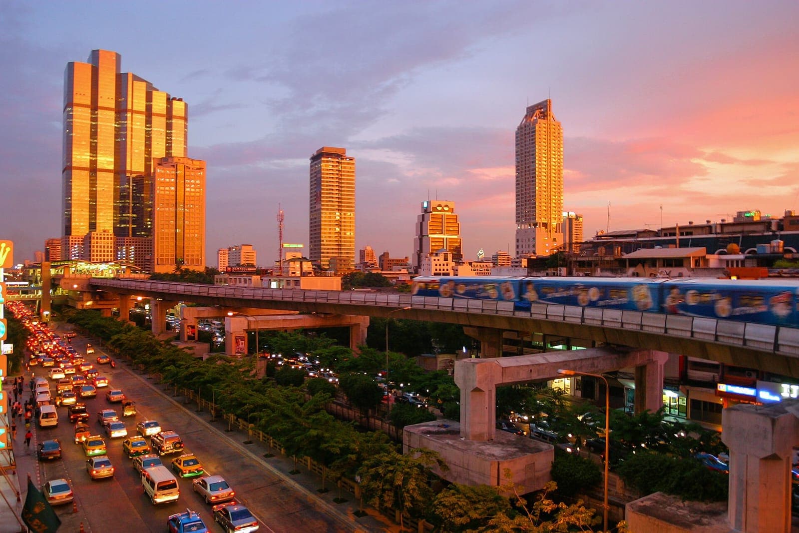 BTS Skytrain crossing an elevated track above heavy road traffic in Bangkok at sunset, with high-rise buildings behind