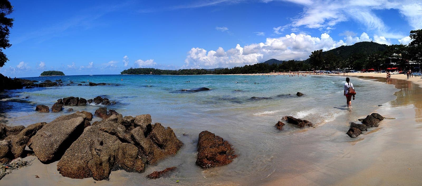 Panorama of Kata Beach in Phuket with rocks in the foreground, turquoise water and a forested headland